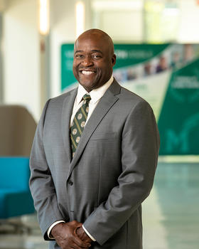 Gregory Washington portrait. He wears a grey three-button suit, and green and gold GMU necktie. He stands in the atrium of Peterson Hall on the Fairfax campus of George Mason University, USA. 