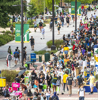 A crowd of students walks through campus, crowding the sidewalks. The photo was taken from an angle that has us looking down towards the students somewhat.